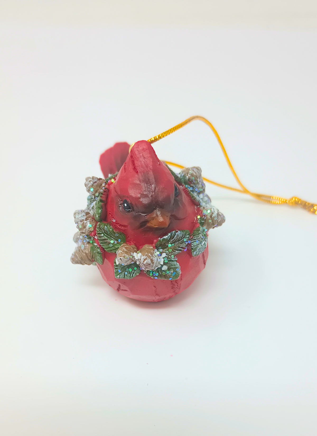 Red cardinal ornament wearing a glittery green wreath with pinecones on a white background.