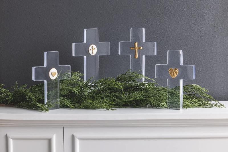 Four clear resin crosses with hand-painted gold icons on a mantel with greenery.