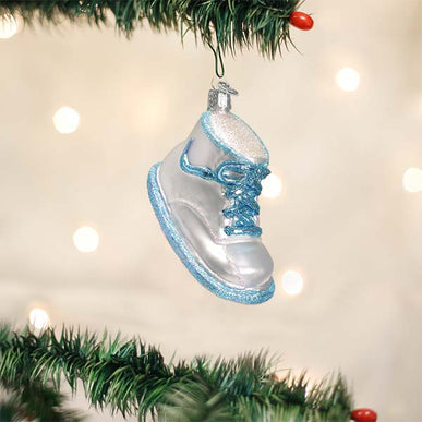 A glittery white and blue glass baby shoe ornament hanging from a Christmas tree branch.