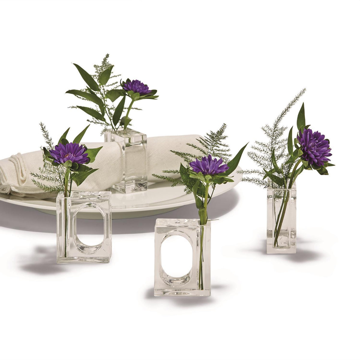 Set of four clear acrylic napkin ring flower holders with purple flowers on a white table setting.