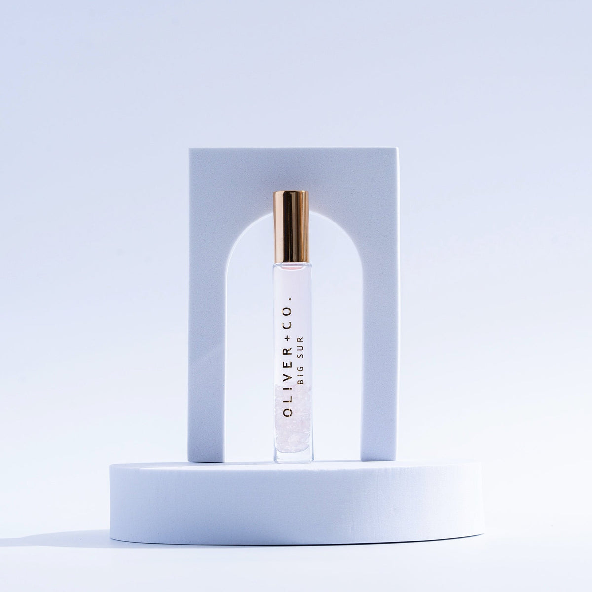 Oliver + Co. Big Sur roll-on perfume stick with a gold cap on a white geometric pedestal.