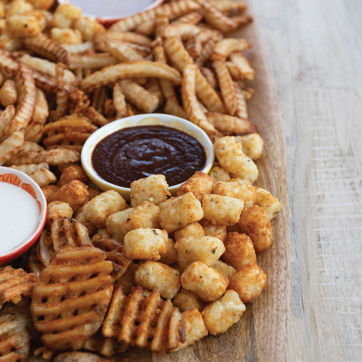 A small bowl of dark Stout BBQ Sauce on a wooden platter surrounded by tater tots and fries.