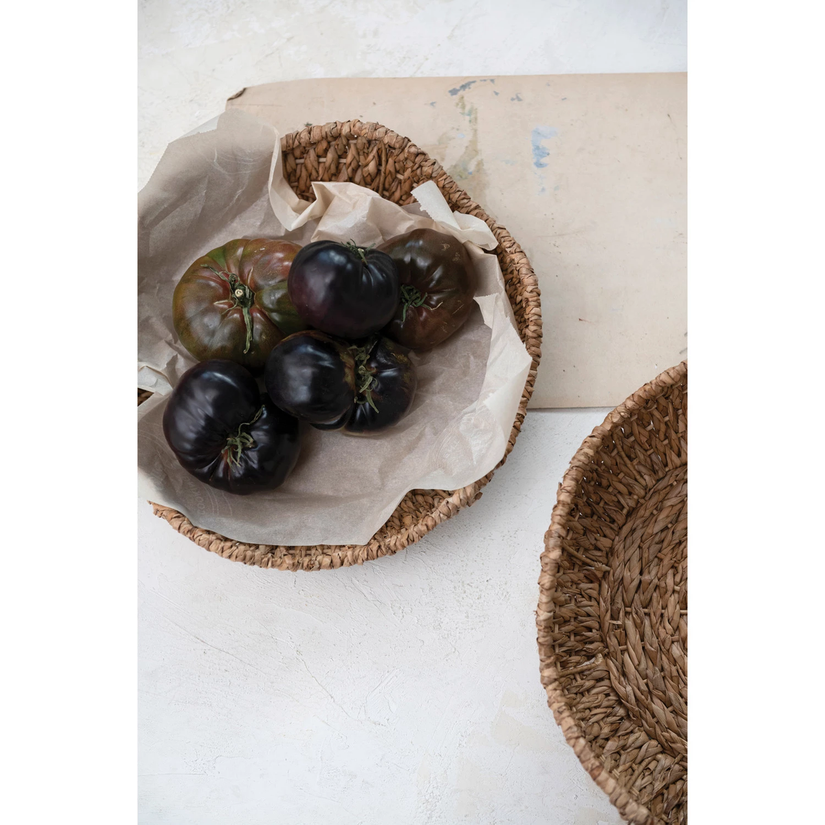 Round braided bankuan bowl with scalloped edges holding dark heirloom tomatoes on parchment paper.