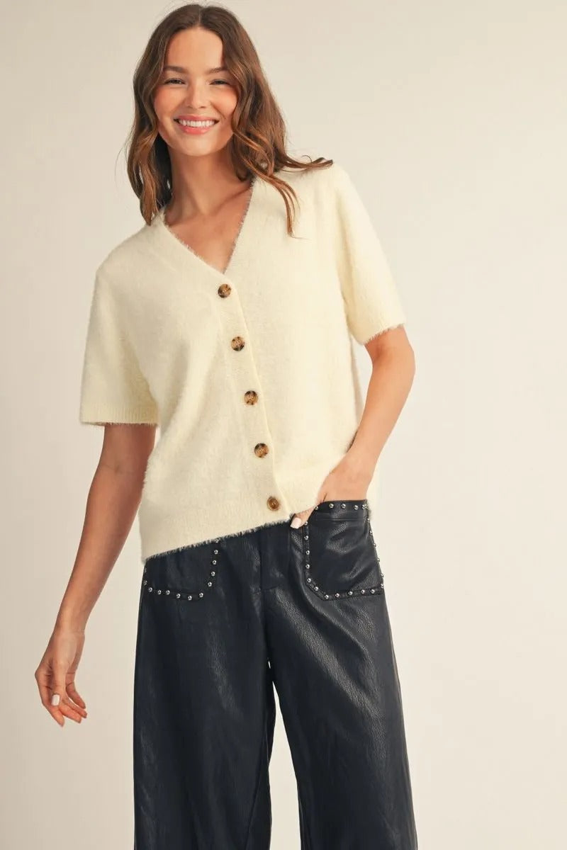 Woman wearing the Aurora short sleeve, cream fuzzy button-down cardigan with black studded pants.