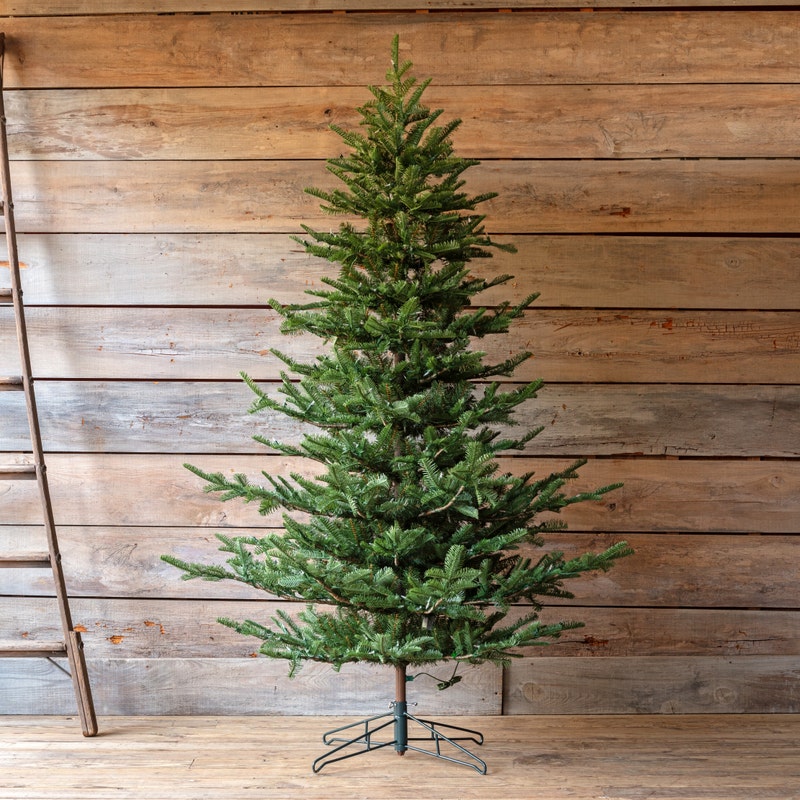Grand Noble 7.5' artificial Christmas tree with green needles on a metal stand against wood walls.