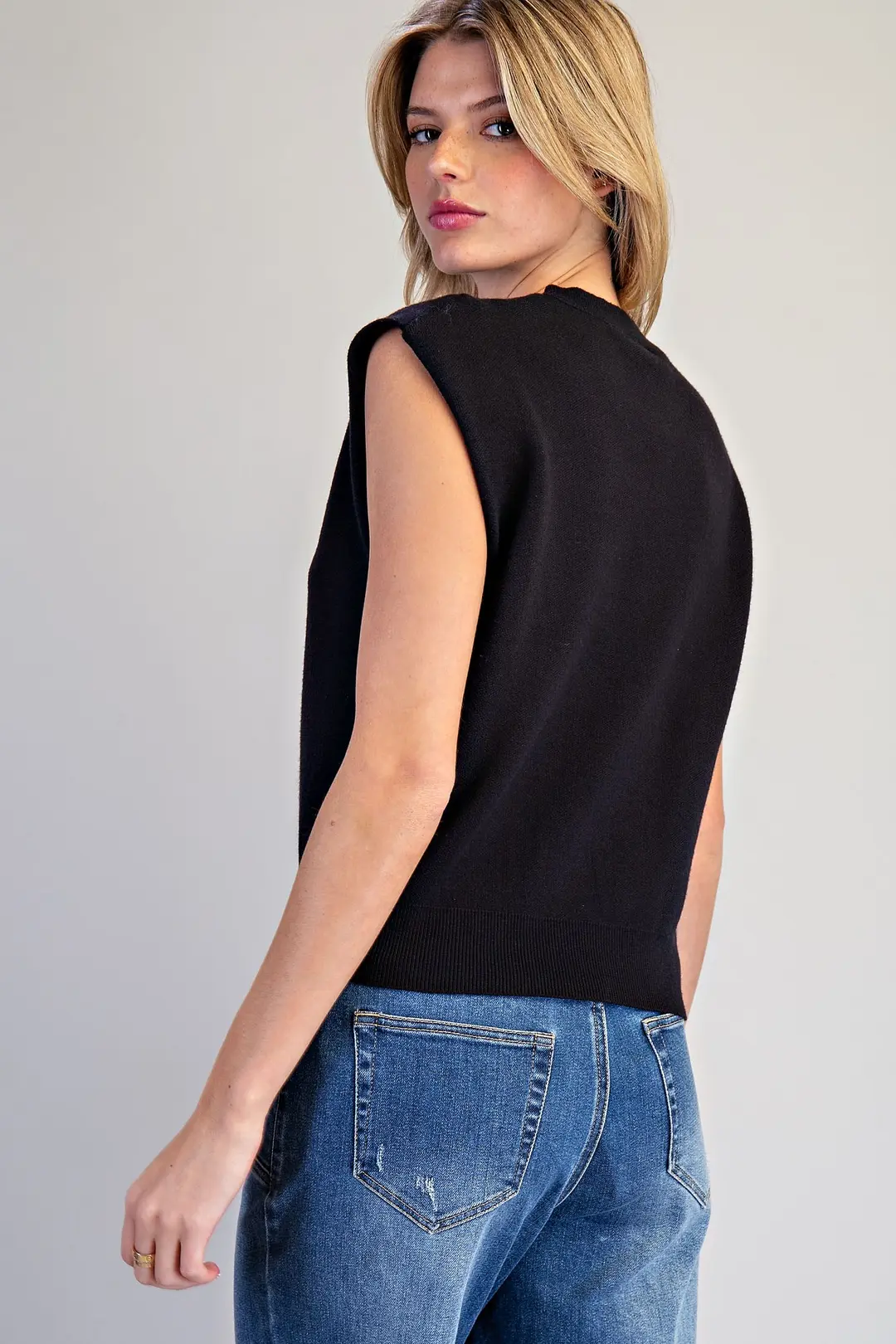 Back view of a woman wearing a black sleeveless knit top with extended shoulders and blue jeans.