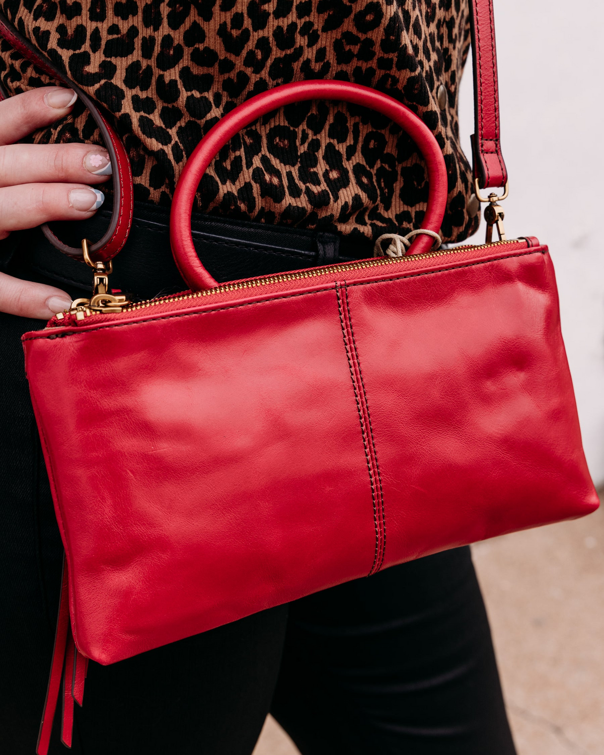 A person wearing a leopard print top holds a red leather Sable Crossbody bag with gold hardware.