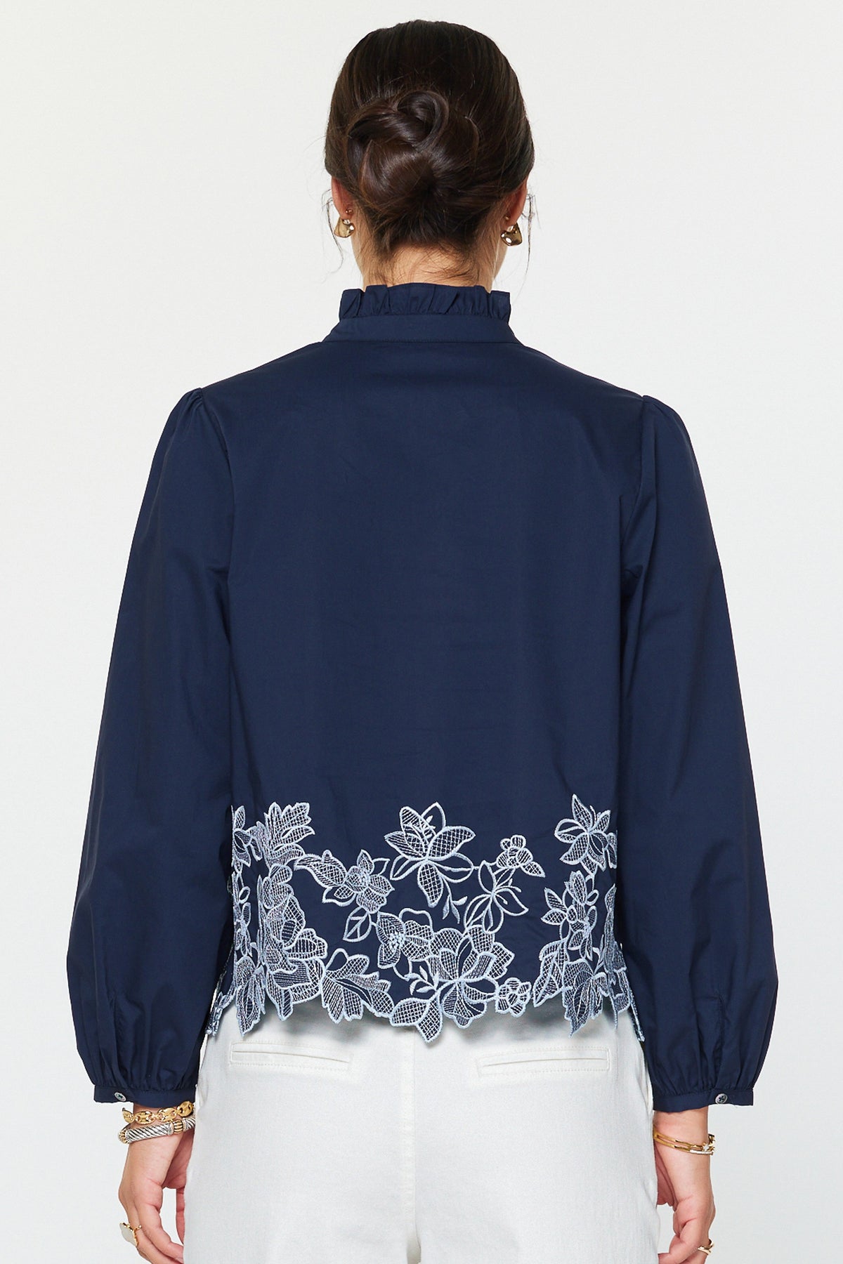 Lucy embroidered long sleeve blouse in cotton poplin with ruffle stand collar, button front closure, puff sleeves, and floral hem embroidery.