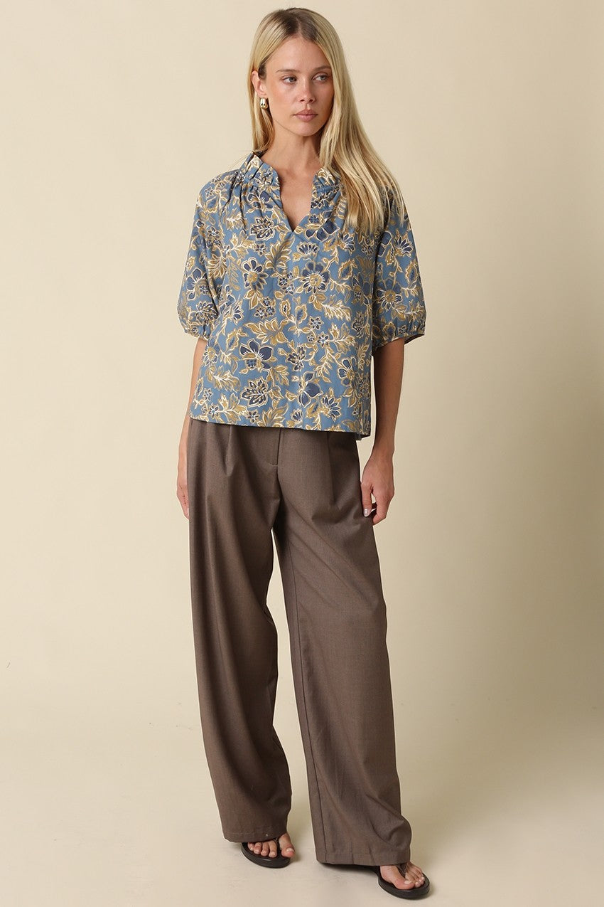 Woman wearing a blue Marina Floral Puff Sleeve Blouse with a ruffle split neck and brown trousers.