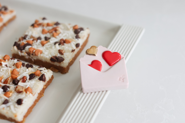 Pink envelope-shaped Love Notes mini with red and gold hearts next to coconut dessert bars.