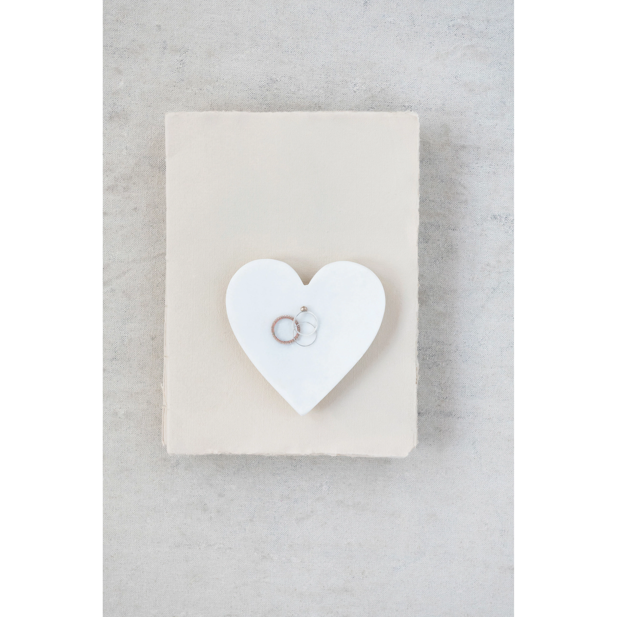 White marble heart-shaped dish holding two rings on top of a cream-colored book.