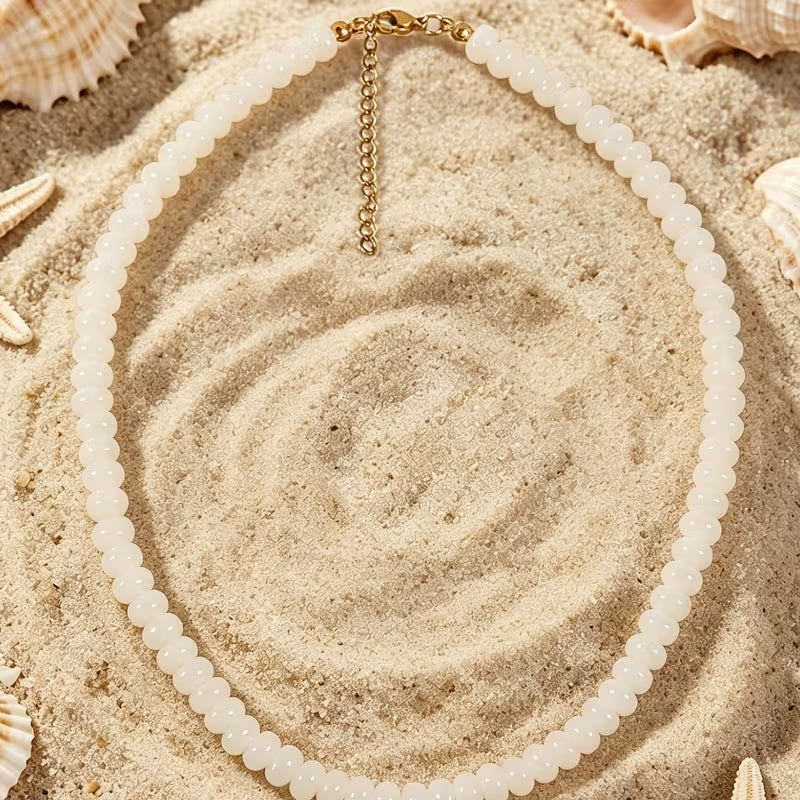 White glass beaded necklace with a gold-tone lobster clasp and extender chain on sand.