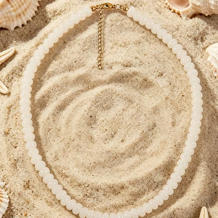 White glass beaded necklace with a gold-tone lobster clasp and extender chain on sand.