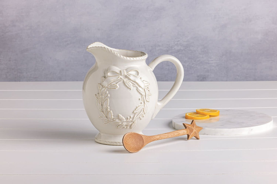 White milk glazed stoneware Christmas pitcher with textured relief details holding a wooden star spoon.