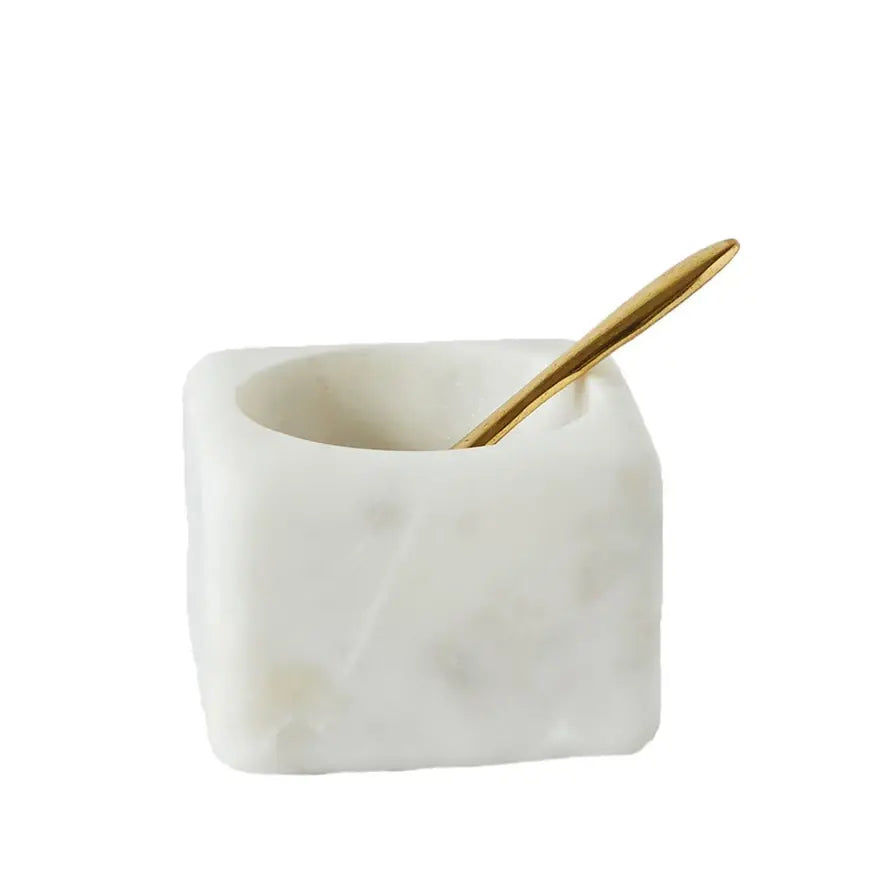 Square white marble bowl with round opening containing a brass spoon