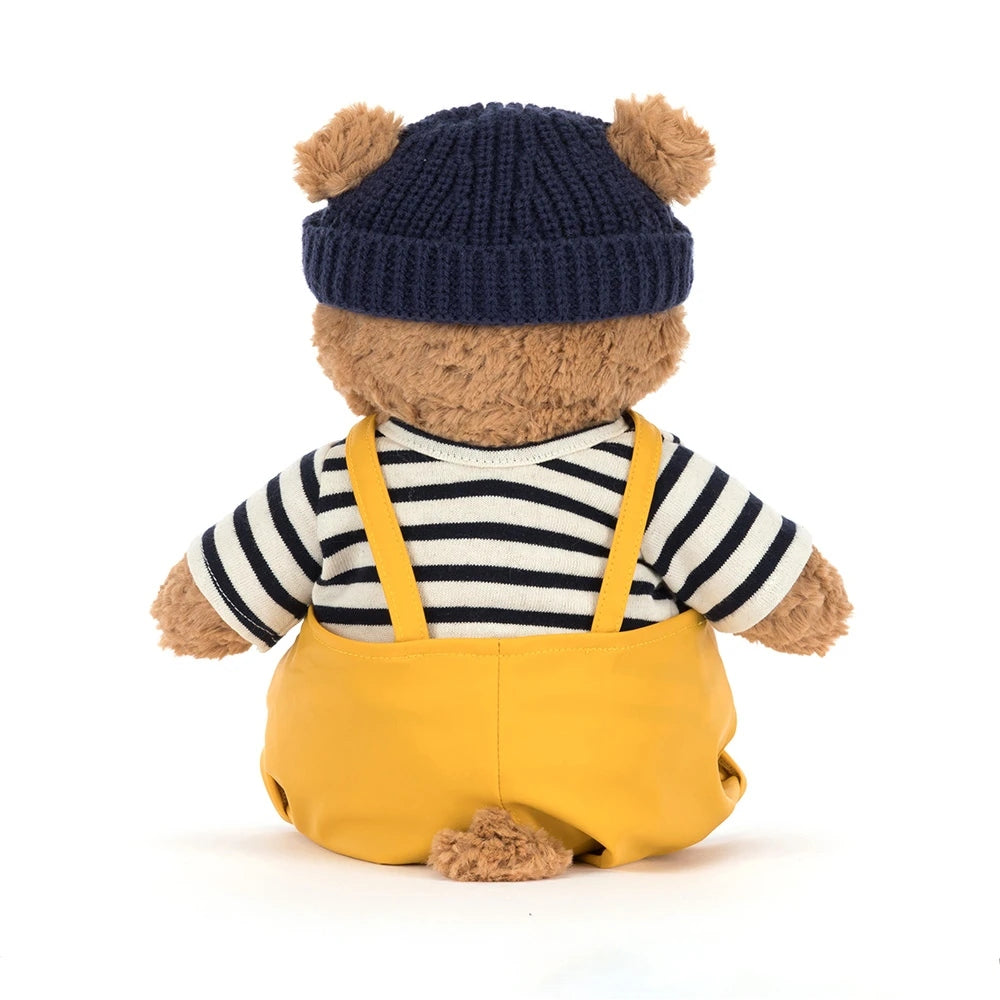 Back view of a brown plush bear wearing a navy beanie, striped shirt, and yellow overalls.