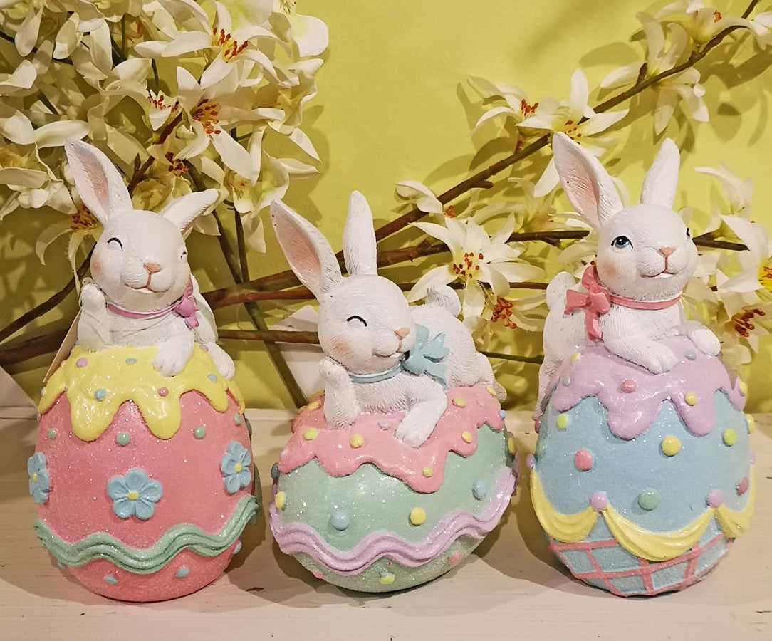 Three white Easter bunny figurines sitting on glittery, pastel-colored eggs with decorative icing and flower accents.