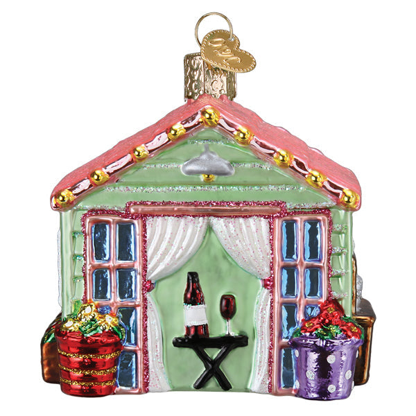 Glass She Shed Christmas ornament with a pink roof, green walls, wine bottle, and floral planters.