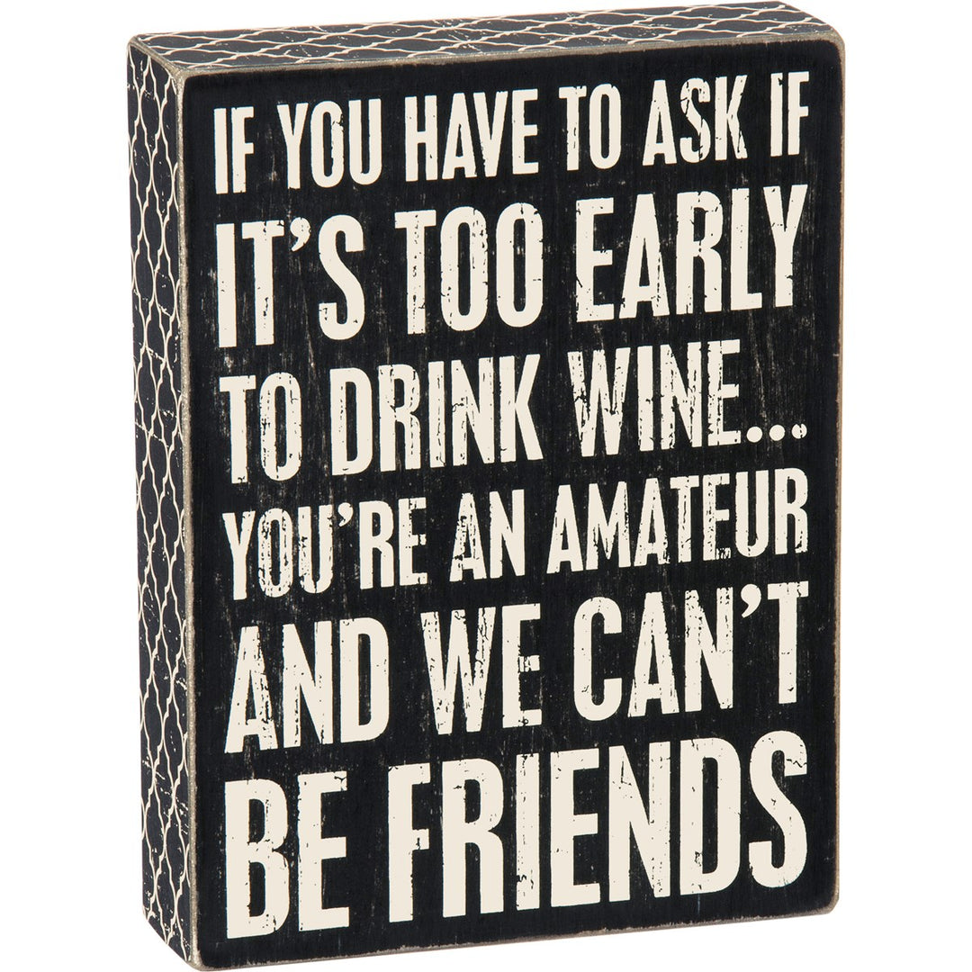 Black wooden box sign with white text that reads 'IF YOU HAVE TO ASK IF IT'S TOO EARLY TO DRINK WINE... YOU'RE AN AMATEUR AND WE CAN'T BE FRIENDS'.