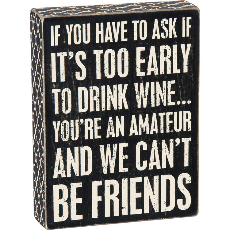 Black wooden box sign with white text that reads 'IF YOU HAVE TO ASK IF IT'S TOO EARLY TO DRINK WINE... YOU'RE AN AMATEUR AND WE CAN'T BE FRIENDS'.