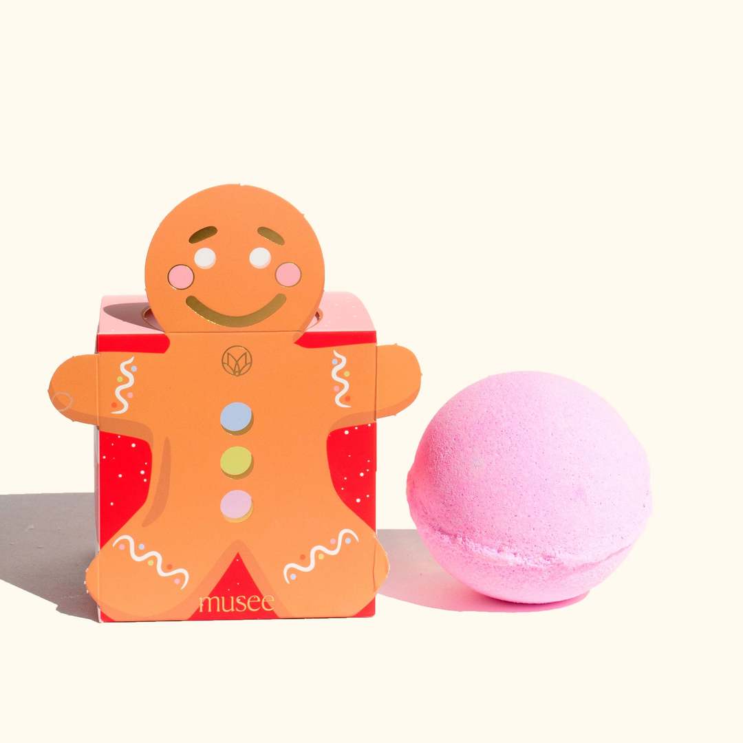 Gingerbread Bath Balm in a gingerbread man shaped box next to a round pink bath bomb.
