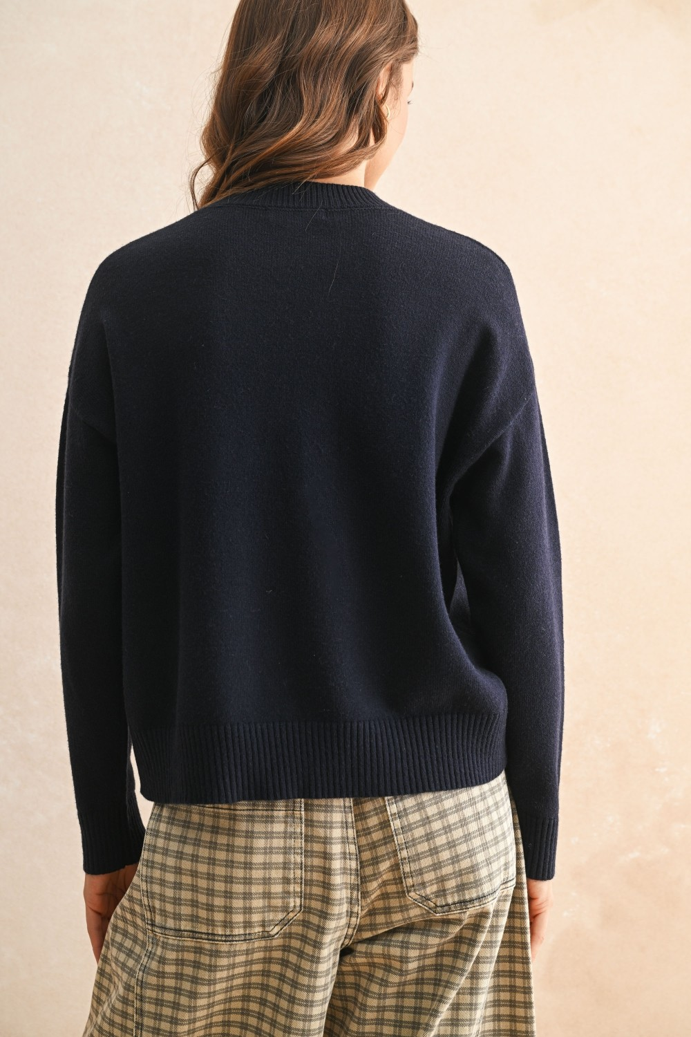 Back view of a woman wearing a navy blue relaxed-fit sweater and plaid trousers.