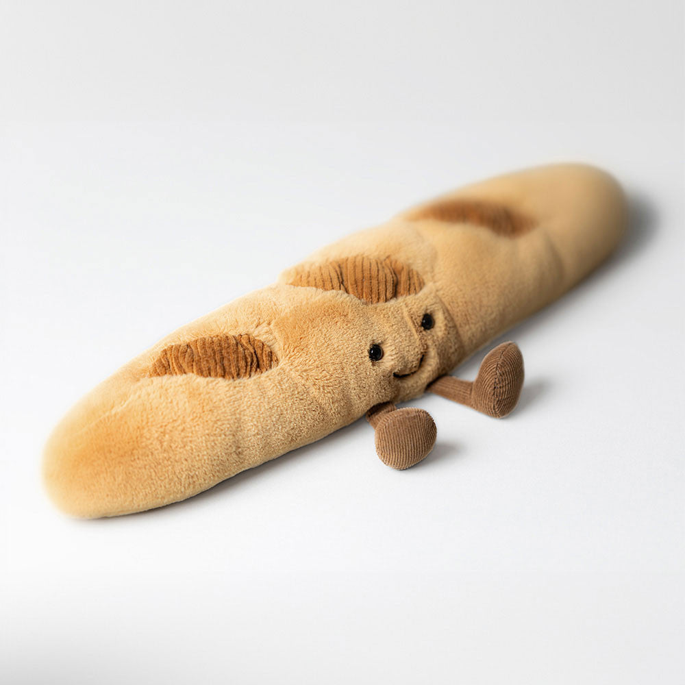 Amuseables Baguette plush toy with a smiling face and cordy legs sitting on a white background.