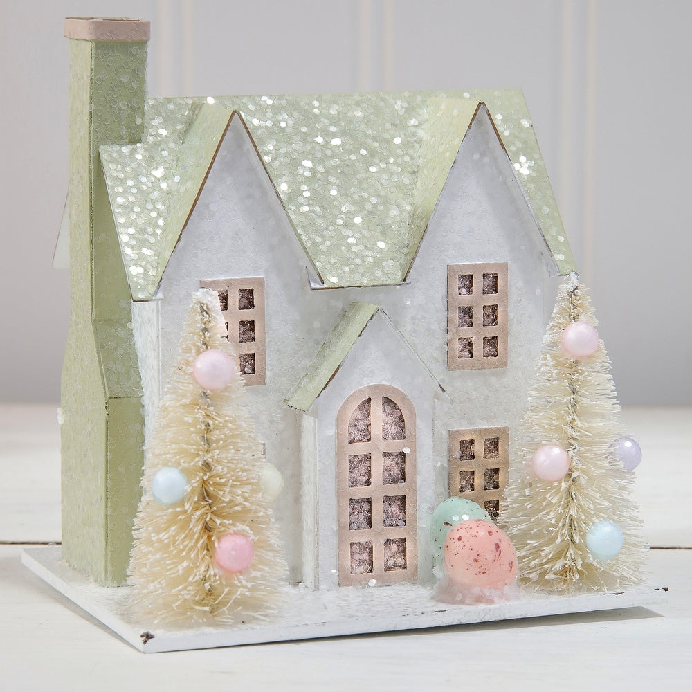 Cardboard Easter house with soft LED lights, sugared finish, and decorative trees in a spring pattern.