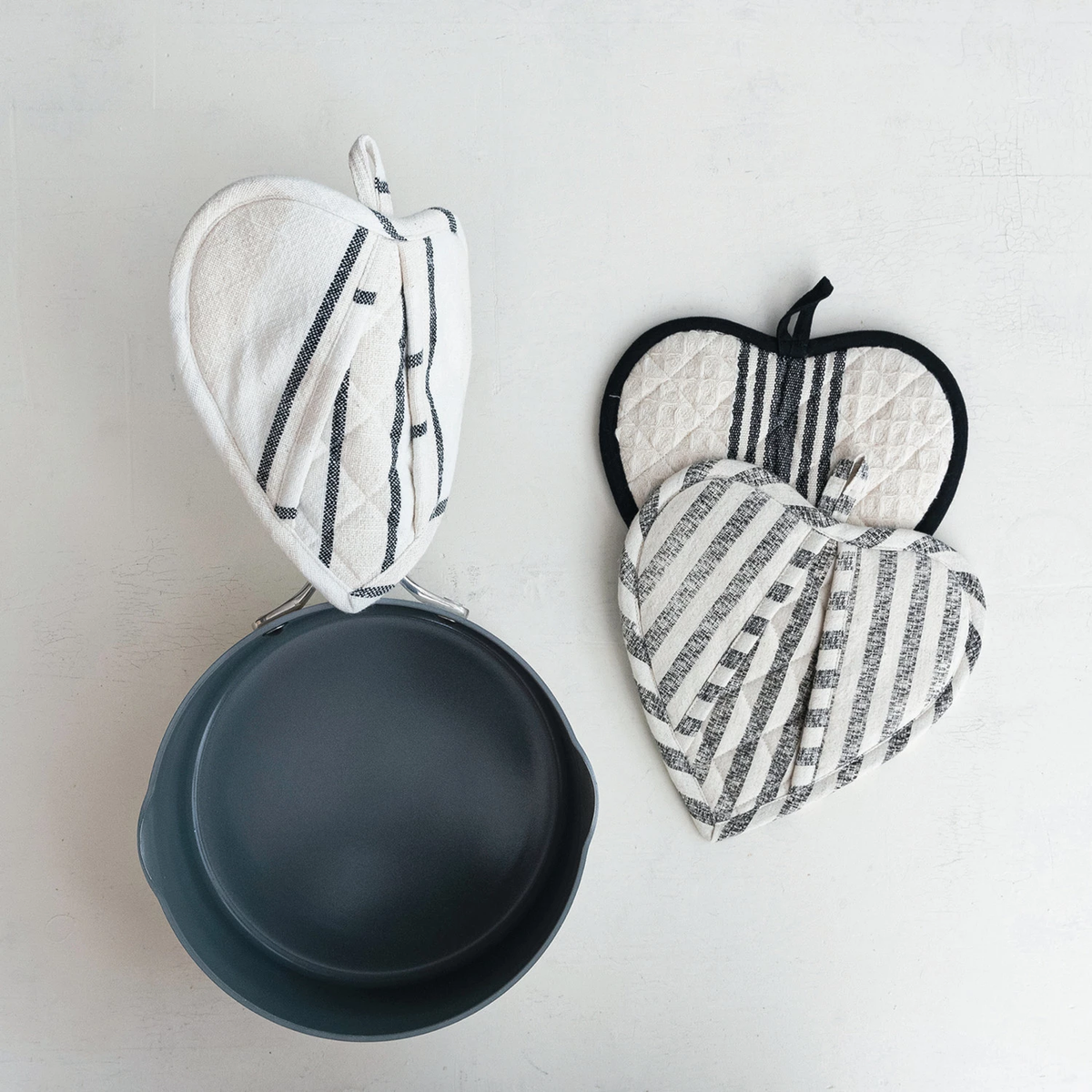 Three cotton quilted heart-shaped pot holders in striped patterns next to a dark grey pan.