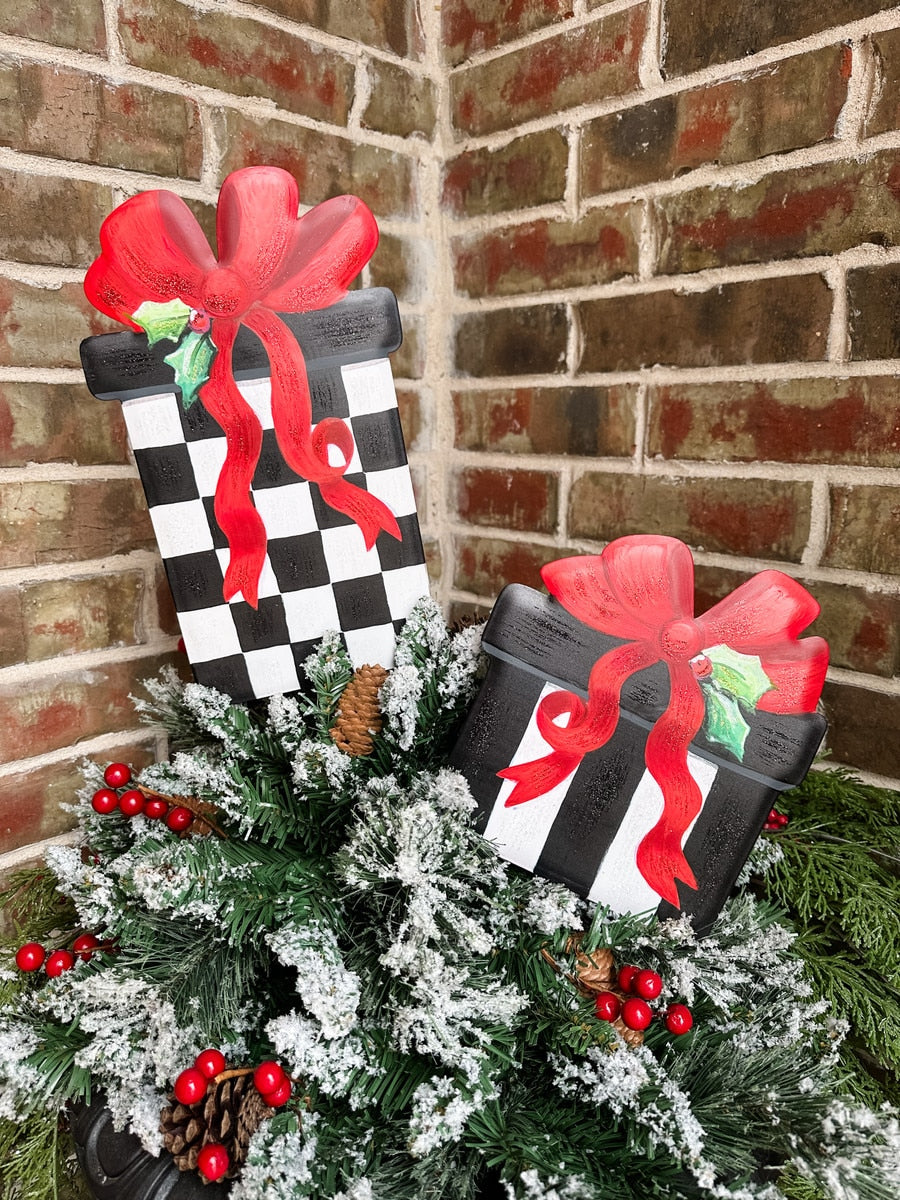 Two decorative gift boxes with red bows, one checkered black and white, the other striped black and white, nestled in faux evergreen branches.