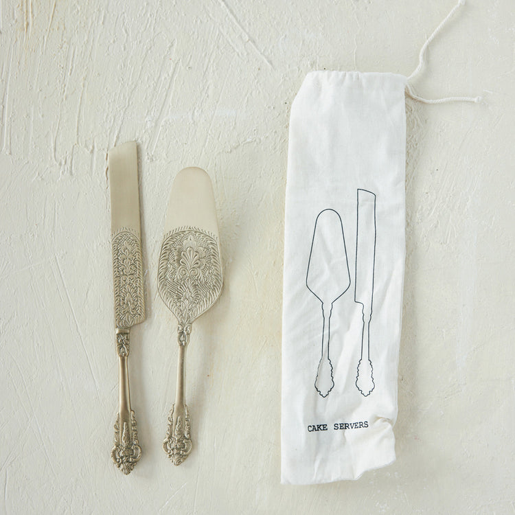 Embossed brass cake knife and server set next to a white printed drawstring storage bag.