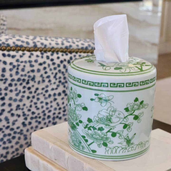 Cylindrical white ceramic tissue vessel with green floral and bird patterns on a stack of books.