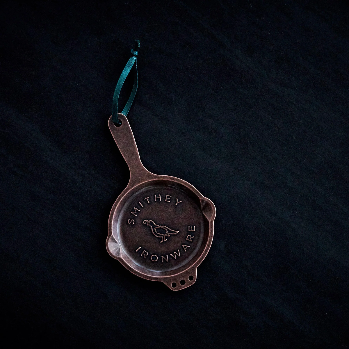 Miniature copper-colored skillet ornament with a quail logo and green ribbon on a dark background.