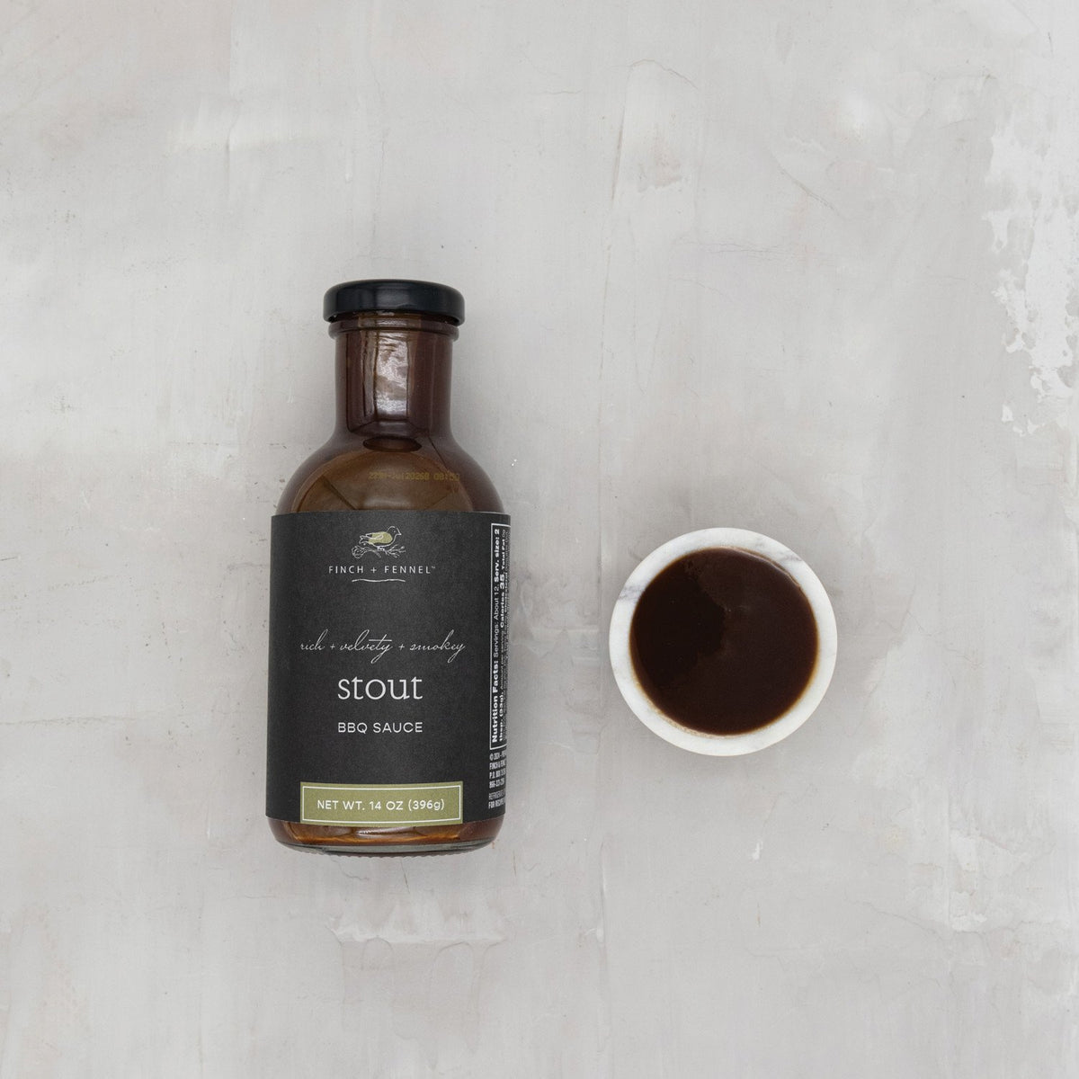 A bottle of Finch + Fennel Stout BBQ Sauce next to a small white bowl filled with the dark sauce.