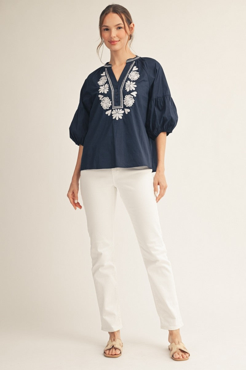 Front view of a white cotton top with balloon sleeves, embroidered floral detail on the chest, notched V-neckline, and front button closure.
