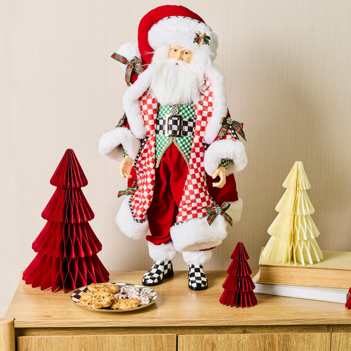 A decorative fabric Santa Claus figure in a checkered coat standing on a table with paper trees.