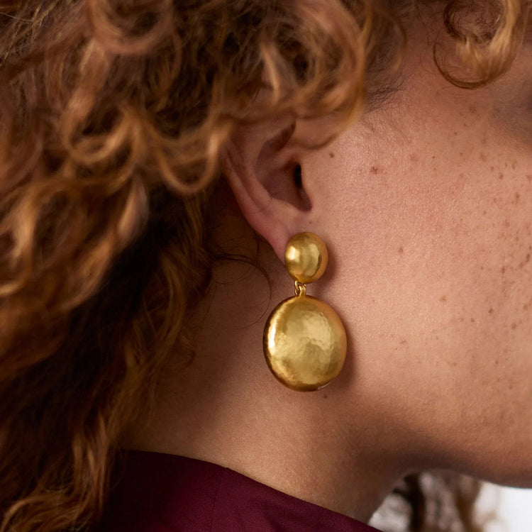 A person with curly hair wearing a Solara Statement Earring in gold with two hammered discs.