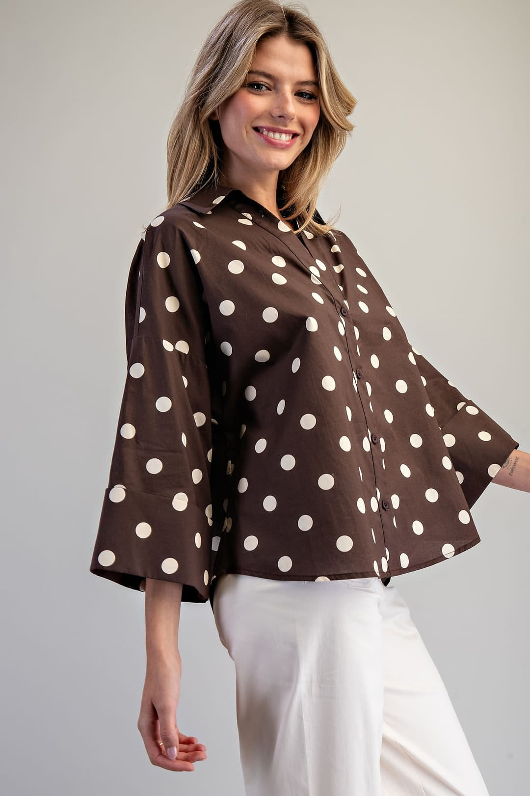 Woman wearing a brown Eliana Button Down Polka Dot Blouse with wide sleeves and white pants.