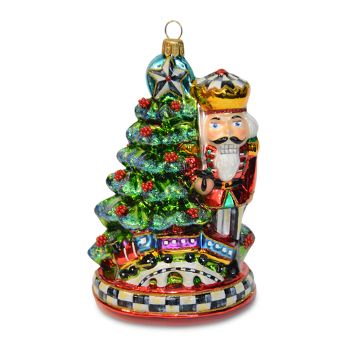 Glass Christmas ornament featuring a nutcracker standing next to a decorated tree with a toy train.