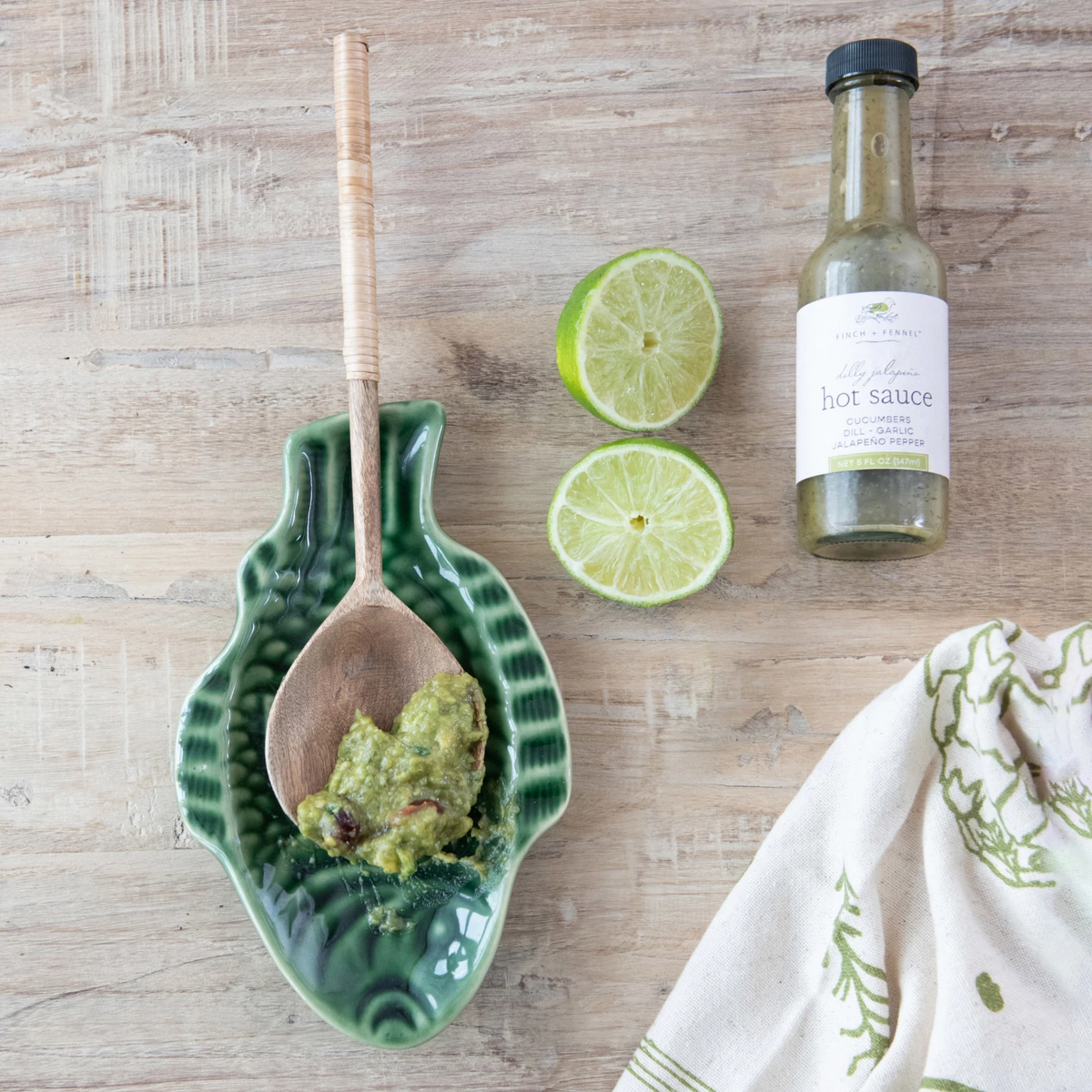 Dilly Jalapeño Hot Sauce bottle next to lime halves and a wooden spoon with green sauce.