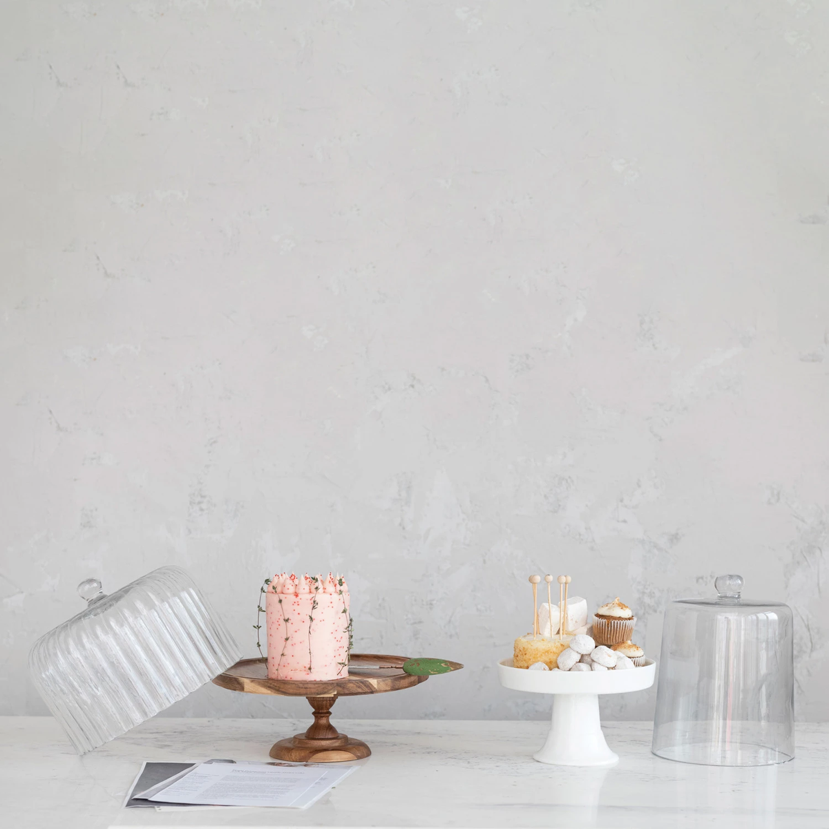 A wooden cake stand with a pink cake and a white ceramic stand with treats and a glass cloche.