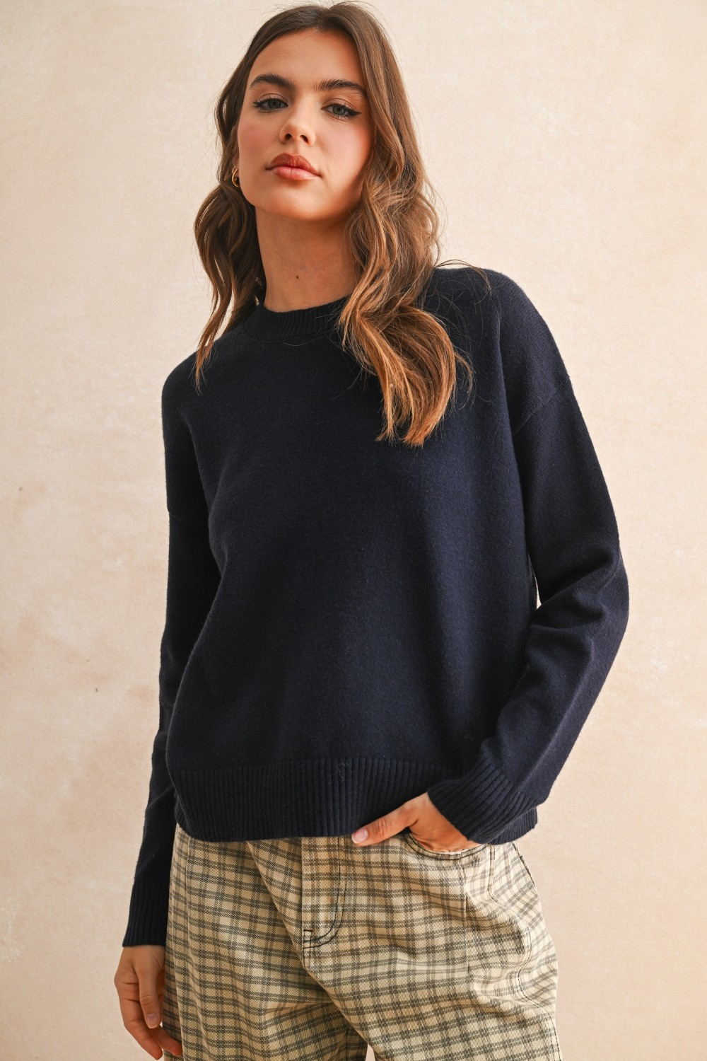 Woman wearing the Ailsa navy blue crew neck sweater with ribbed cuffs and plaid trousers.
