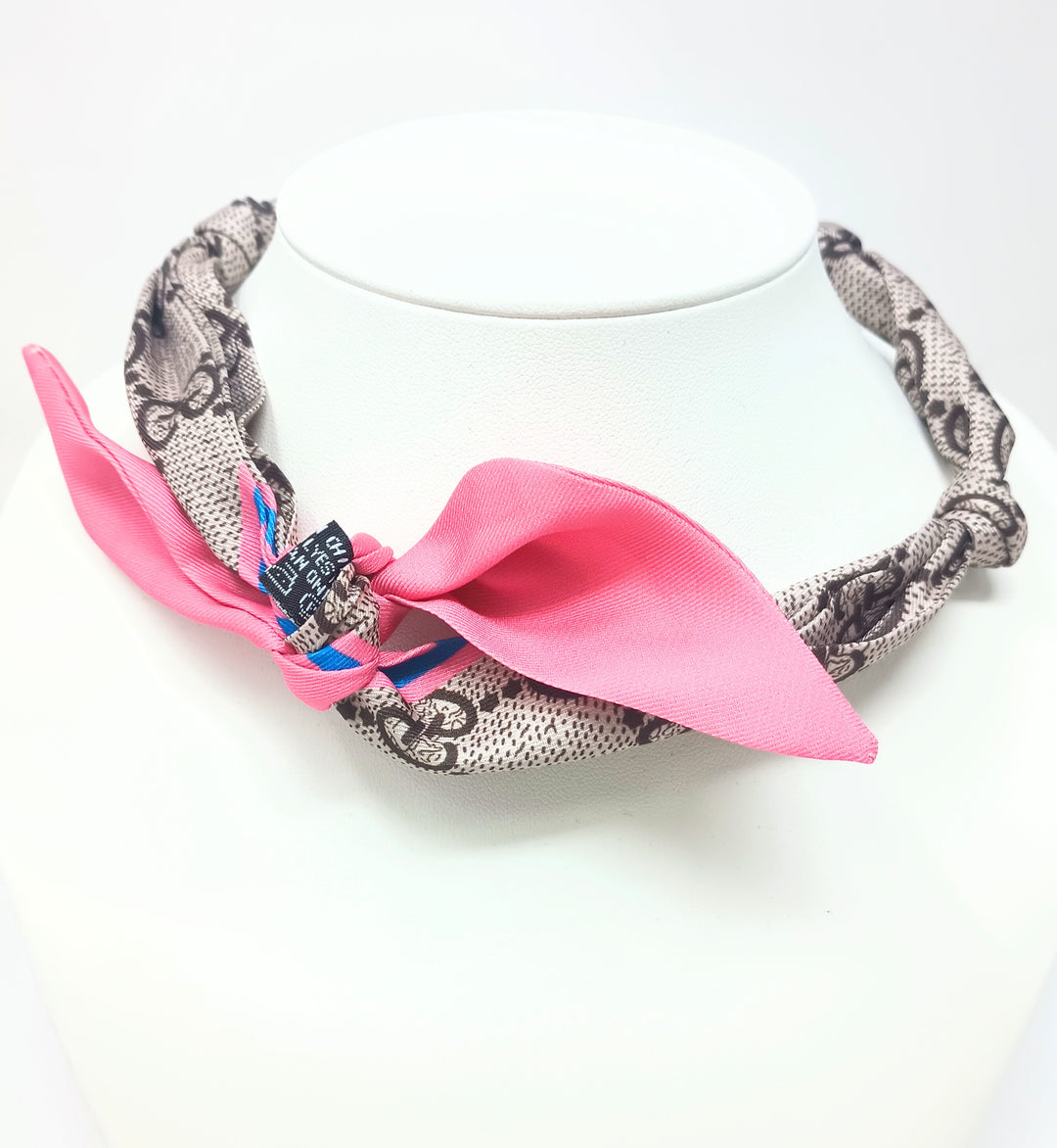 Knotted satin scarf necklace with a tan and black pattern and a bright pink bow on a white display.