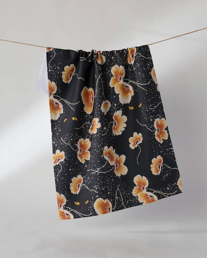 Halloween tea towel with orange flowers and glowing cat eyes on a black starry background.
