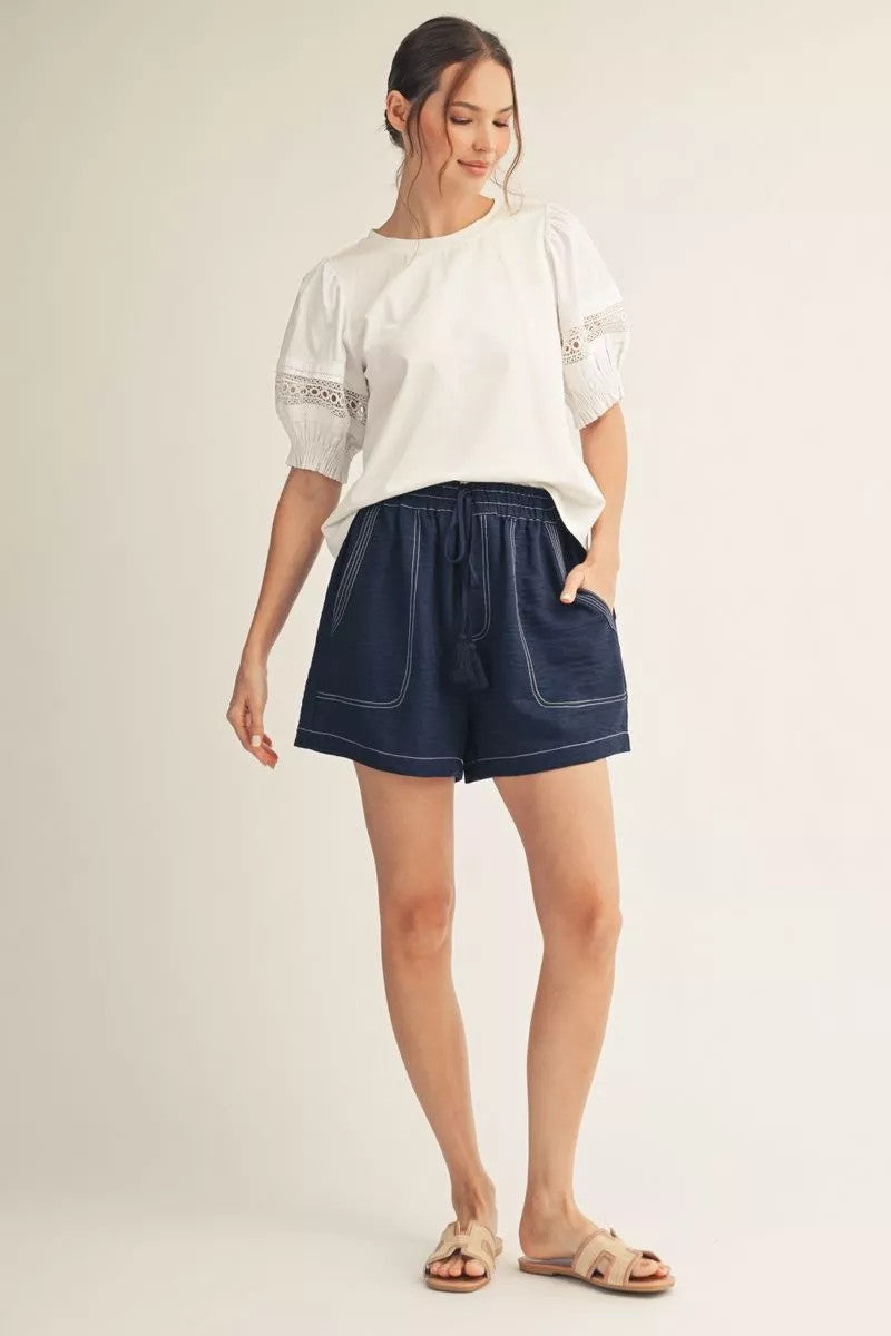 Woman wearing a white Madison short puff sleeve top with lace contrast and navy drawstring shorts.