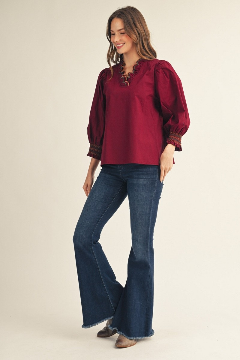 Woman wearing a burgundy Millie top with scalloped V-neckline and flared jeans.