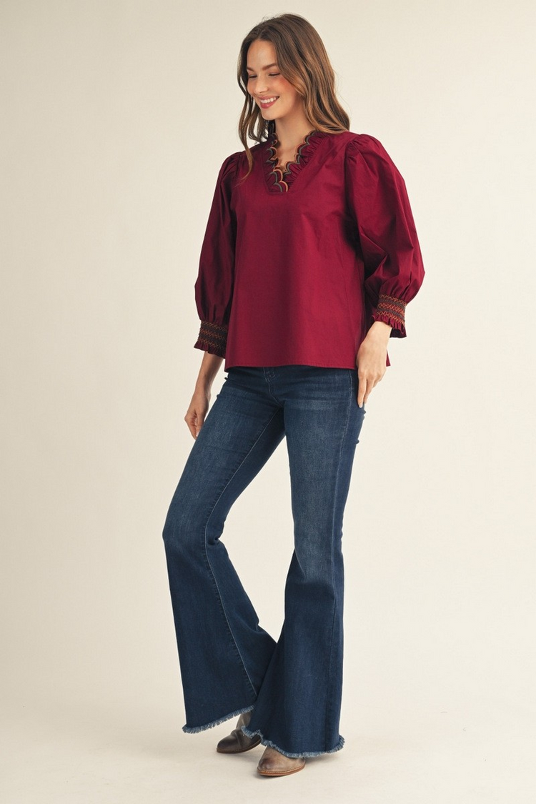 Woman wearing a burgundy Millie top with scalloped V-neckline and flared jeans.