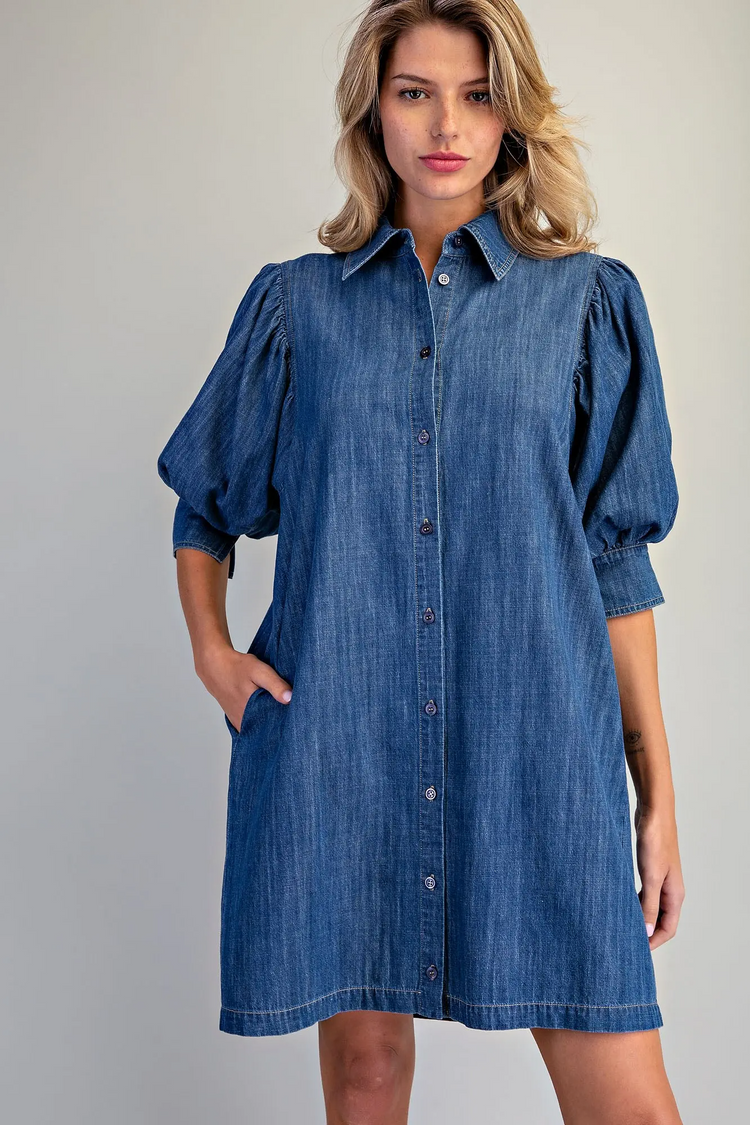 Woman wearing a blue denim button-down mini dress with puff sleeves and side pockets.