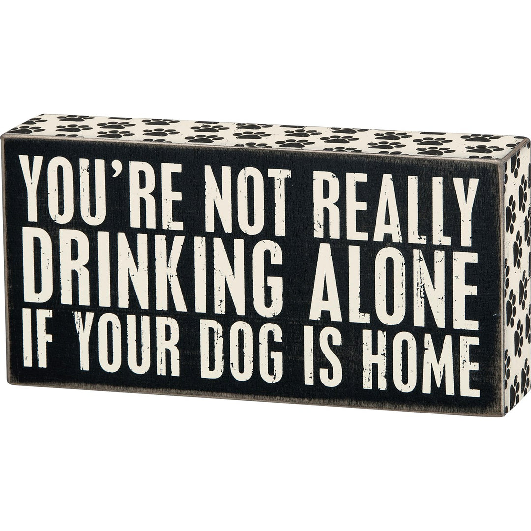 Black wooden box sign with white text reading "YOU'RE NOT REALLY DRINKING ALONE IF YOUR DOG IS HOME". Paw print pattern on top and sides.