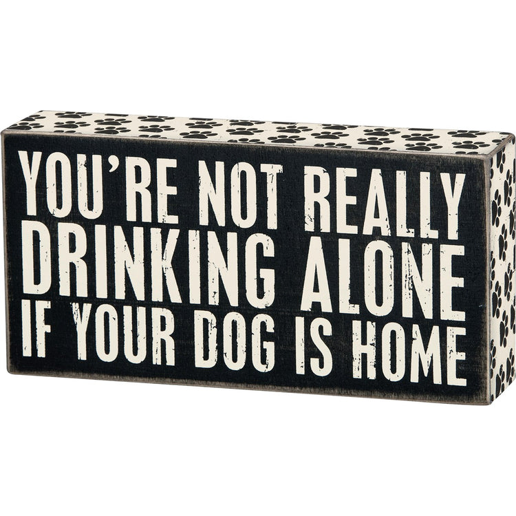 Black wooden box sign with white text reading "YOU'RE NOT REALLY DRINKING ALONE IF YOUR DOG IS HOME". Paw print pattern on top and sides.
